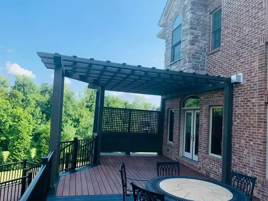 Brick house deck with wooden pergola, black railing, and round patio table overlooking greenery