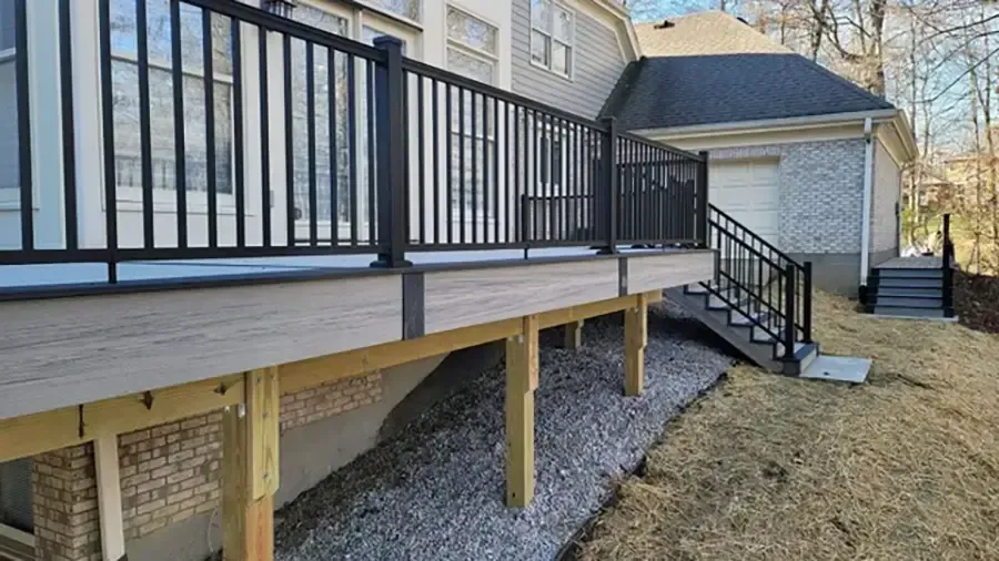 Raised wooden deck with black railing and stairs beside a house, overlooking a gravel yard
