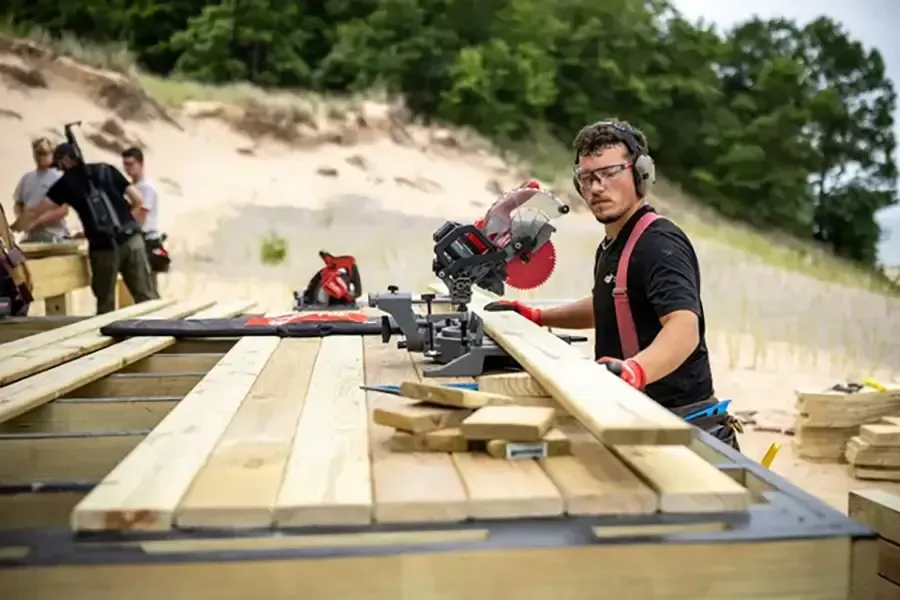 Man using a miter saw to cut wood on an outdoor workbench near sand dunes
