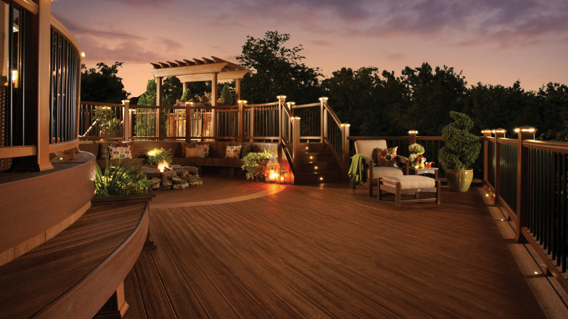 Wooden rooftop deck at sunset with lounge chairs, pergola, plants, and city lights beyond the railing