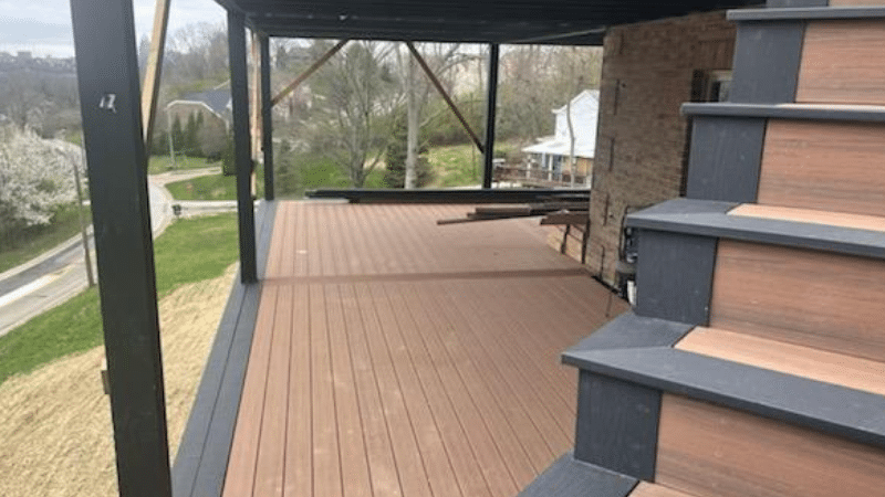 Covered wooden deck with dark railings and steps overlooking a yard and neighborhood street