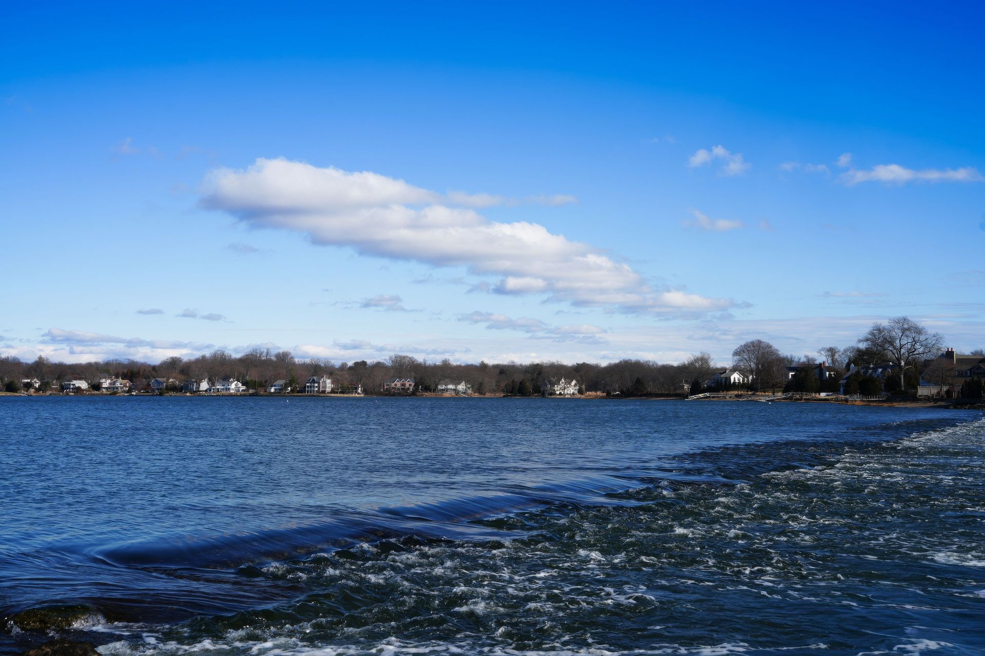 Calm blue lake with a distant shoreline under a bright sky, seen from a rocky shore