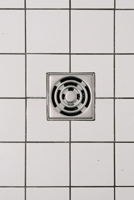 Square metal drain cover centered in a white tiled wall or shower floor.