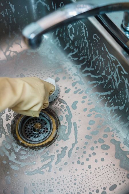 Gloved hand scrubbing a stainless steel sink with soapy water and a sponge