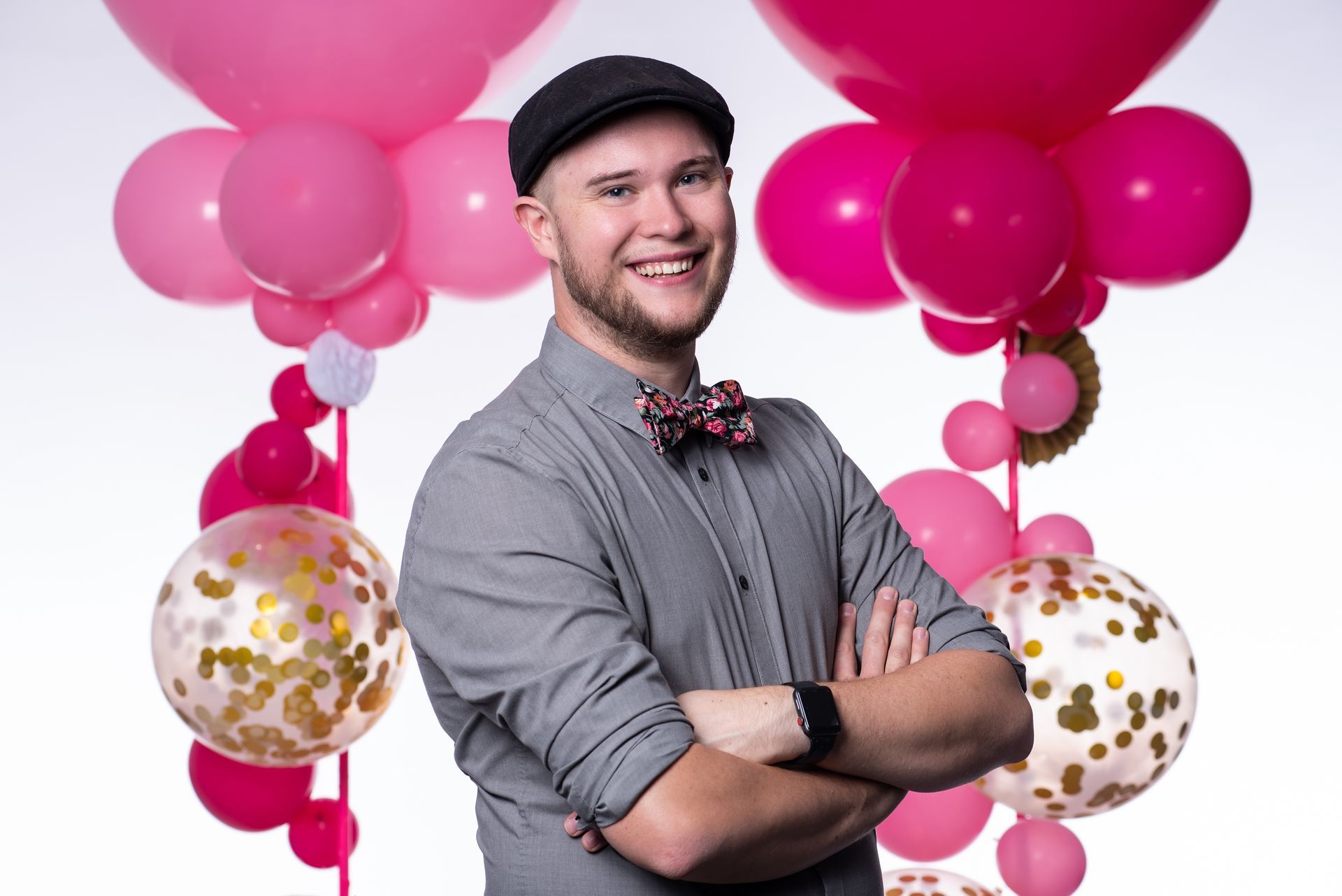 A man in a hat and bow tie is standing in front of pink balloons.