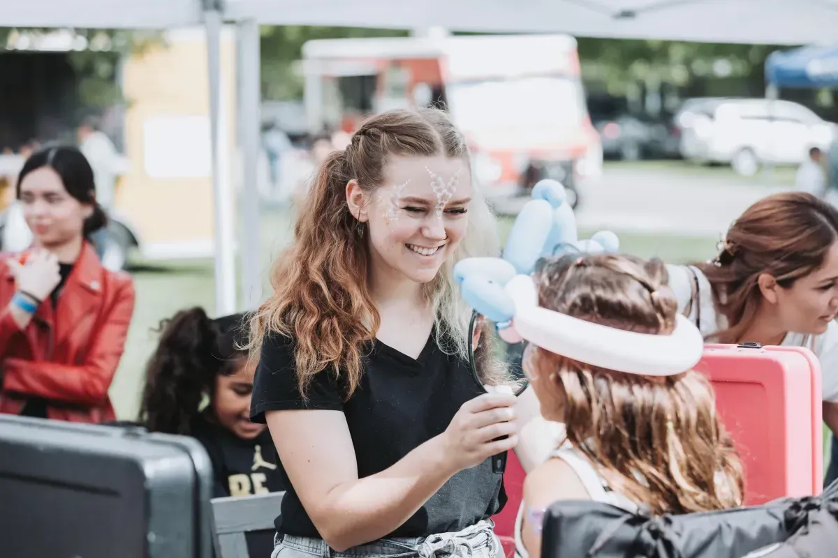 A woman is painting a child 's face with balloons at a festival.