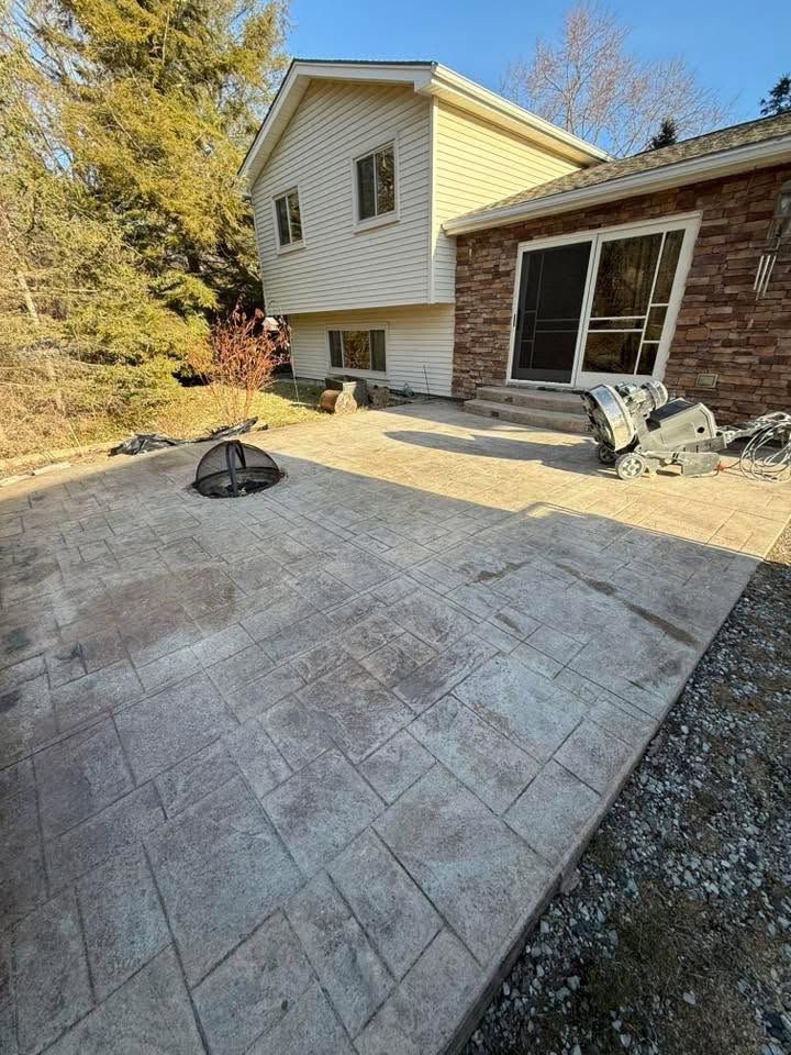 Stamped concrete patio beside a brick house, with a small fire pit and wooded backyard in the background