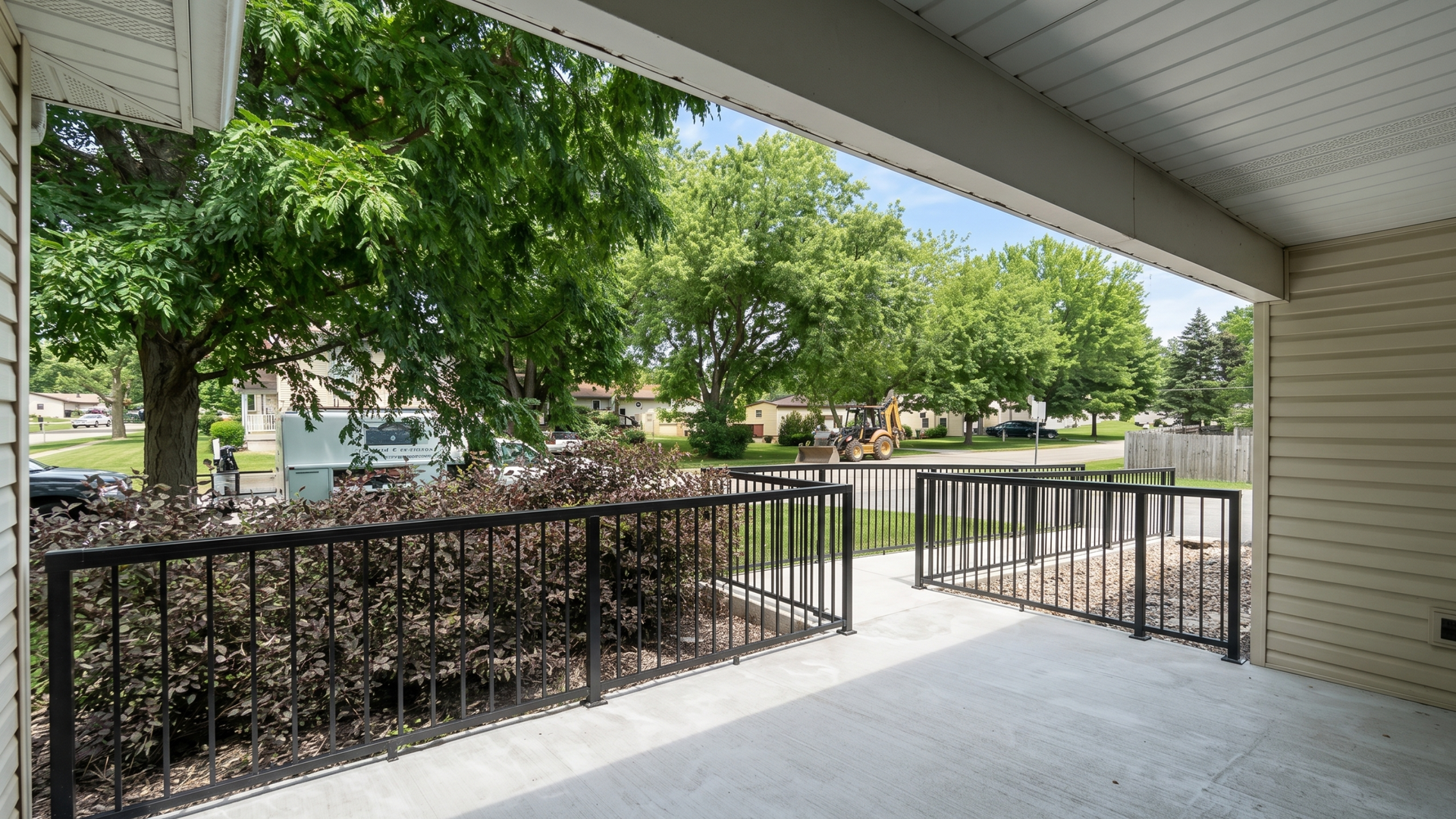 Covered porch with black railing overlooking a green park and sidewalk trees