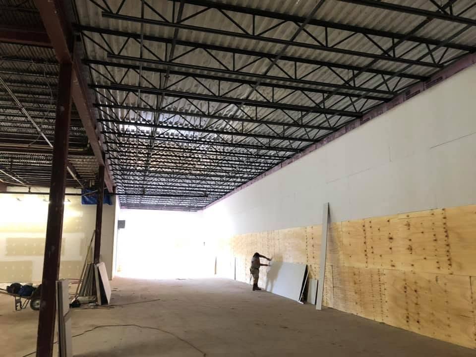 Large warehouse interior with exposed steel roof, white walls, plywood panels, and a person working near the far wall