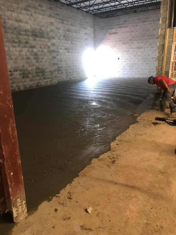 Worker smoothing wet concrete floor inside a warehouse with cinderblock walls