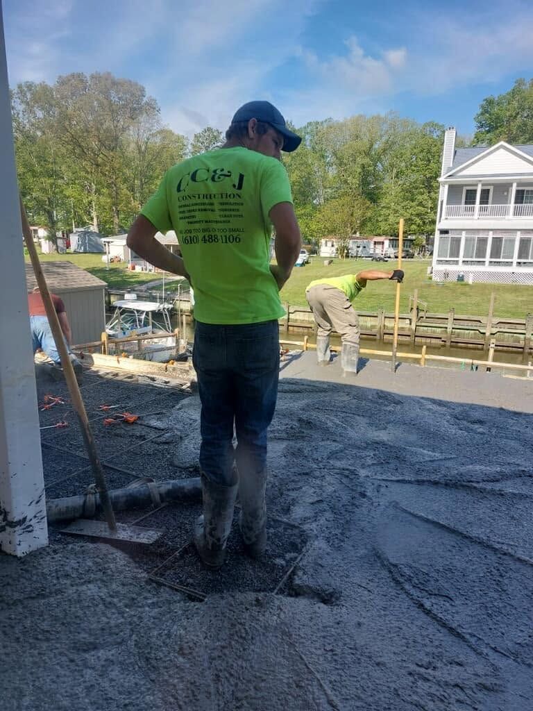 Worker spreading wet concrete on a residential driveway near a house and lake.