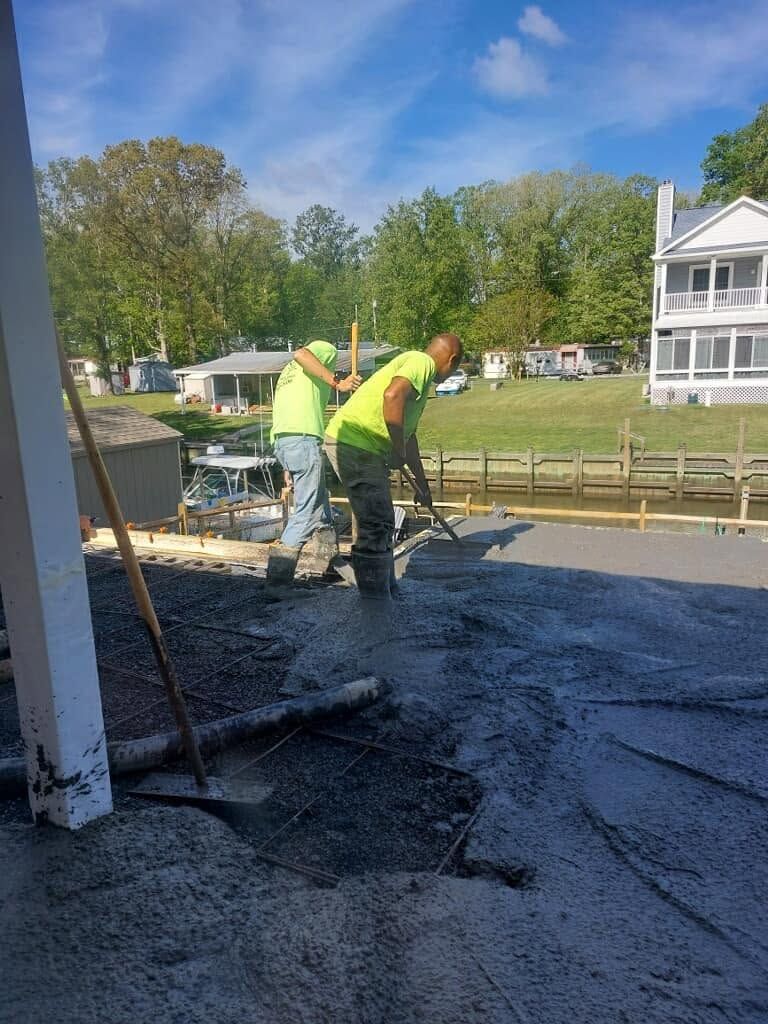 Two workers in neon vests pour concrete on a driveway beside a white house and lake