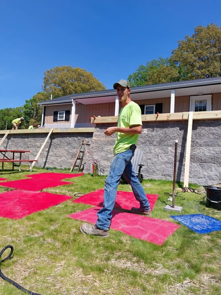Man in neon green shirt and jeans walking across a colorful painted lawn beside a stone wall and house
