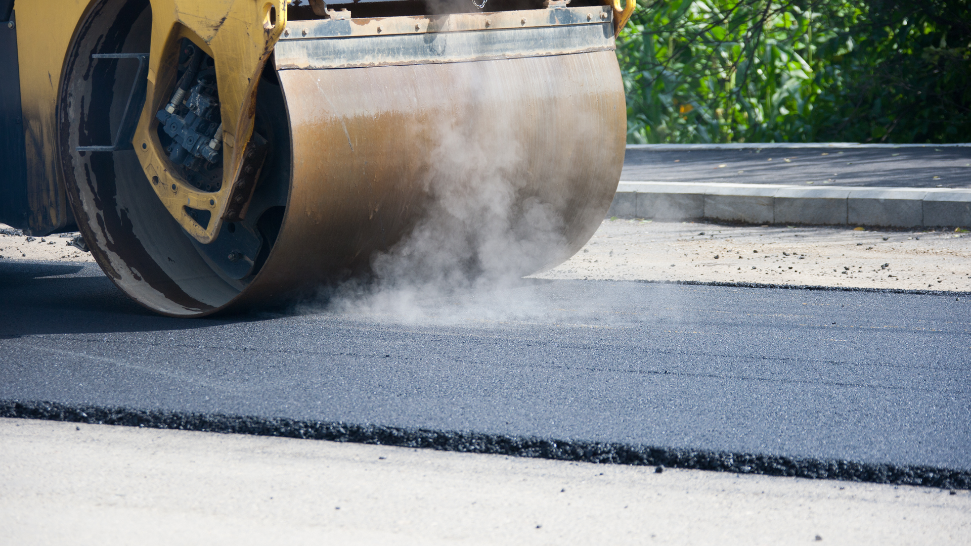 Road roller compacting fresh asphalt on a paved street, with dust rising from the drum.