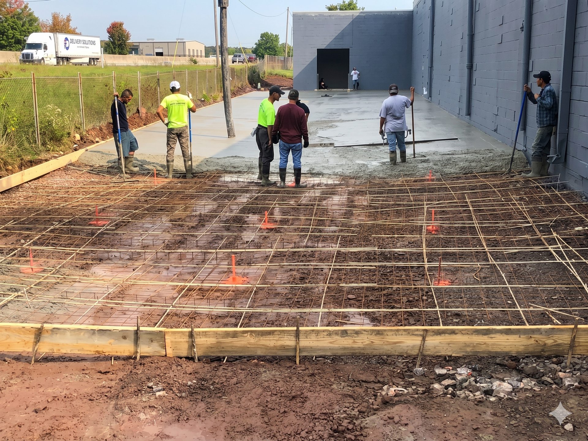 Workers pouring concrete on a rebar foundation beside a gray building at a construction site