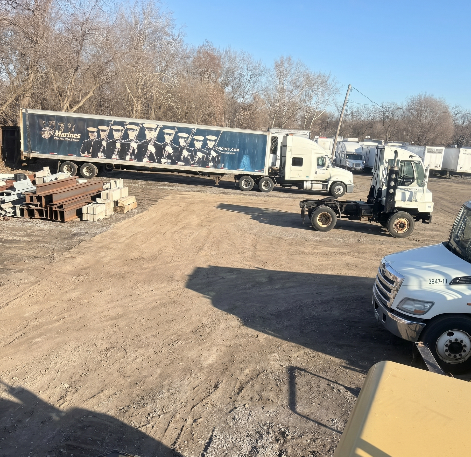 Truck yard with two semi-trucks, a white pickup, and stacked bricks on a dirt lot under bare trees.