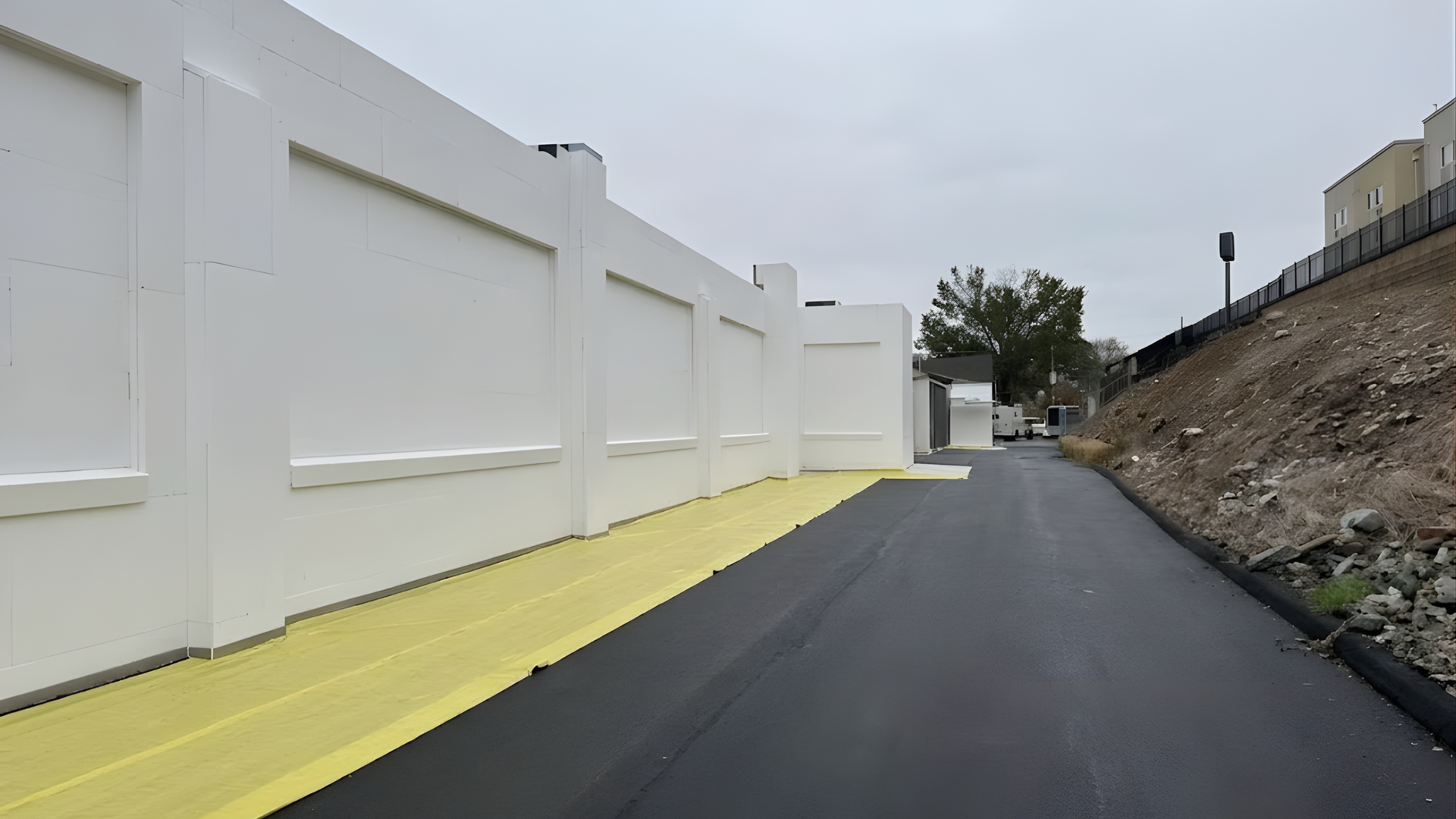Empty paved path beside a white wall and yellow curb, with a rocky slope on the right.
