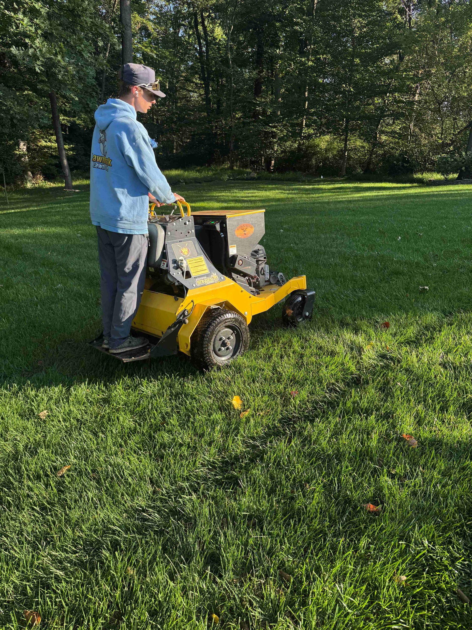 Person operating a yellow lawn mower on a grassy field near trees.