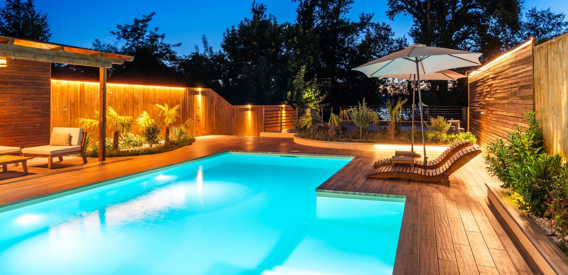 Nighttime view of a swimming pool, surrounded by wooden decking and walls, with lounge chairs and an umbrella.