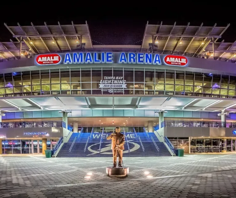 Amalie Arena lit up at night with a statue in front and blue-lit entrance stairs.