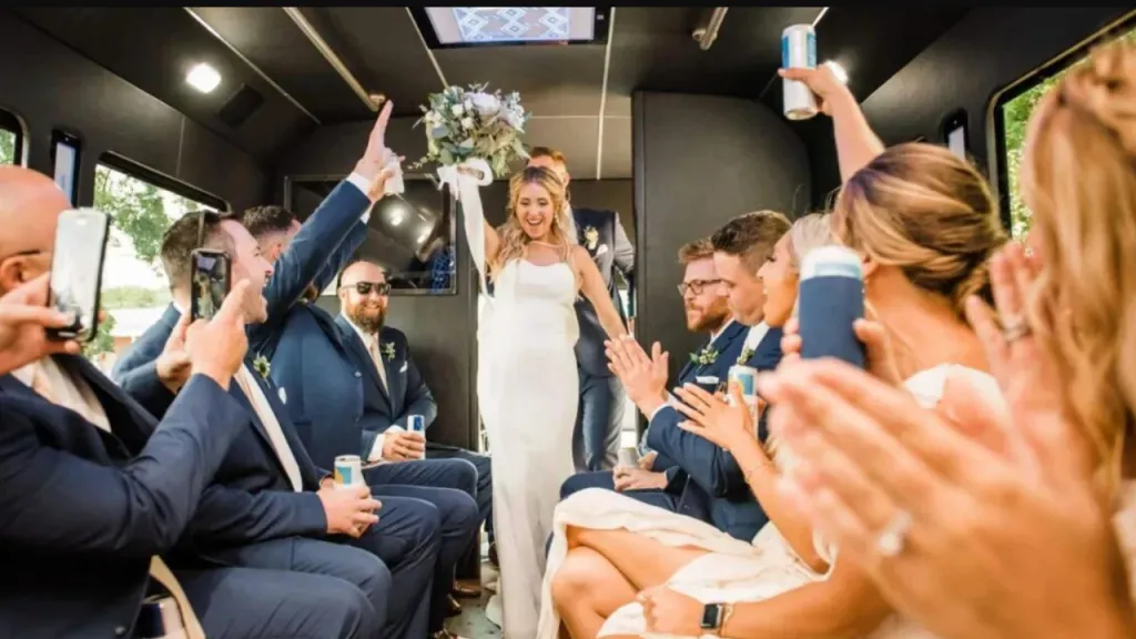 Bride stands in a decorated party bus as seated guests clap and celebrate her.