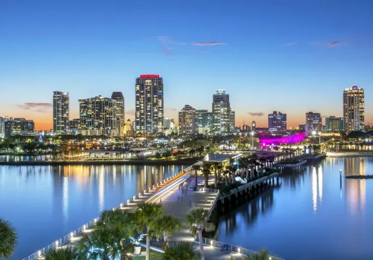 City skyline at dusk reflecting on calm water, with a lit bridge and colorful waterfront lights