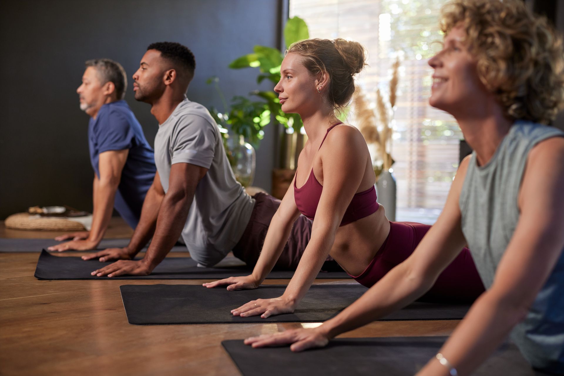 Quattro persone in una lezione di yoga, che eseguono la posizione del cane a testa in su. Sono sedute su dei tappetini in una stanza luminosa.