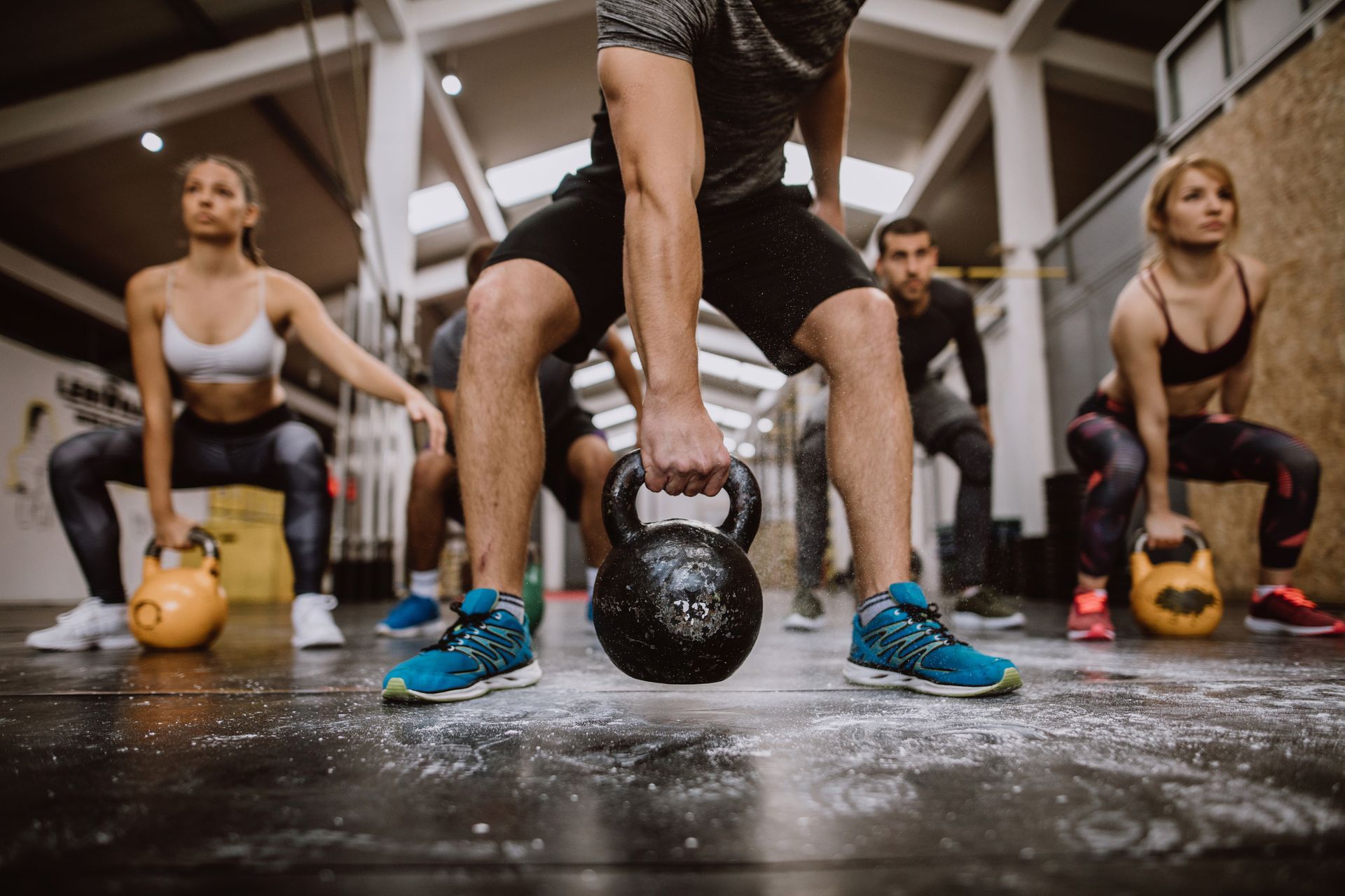 Gruppo di persone che si allenano con i kettlebell in palestra, eseguendo squat, primo piano della persona centrale.