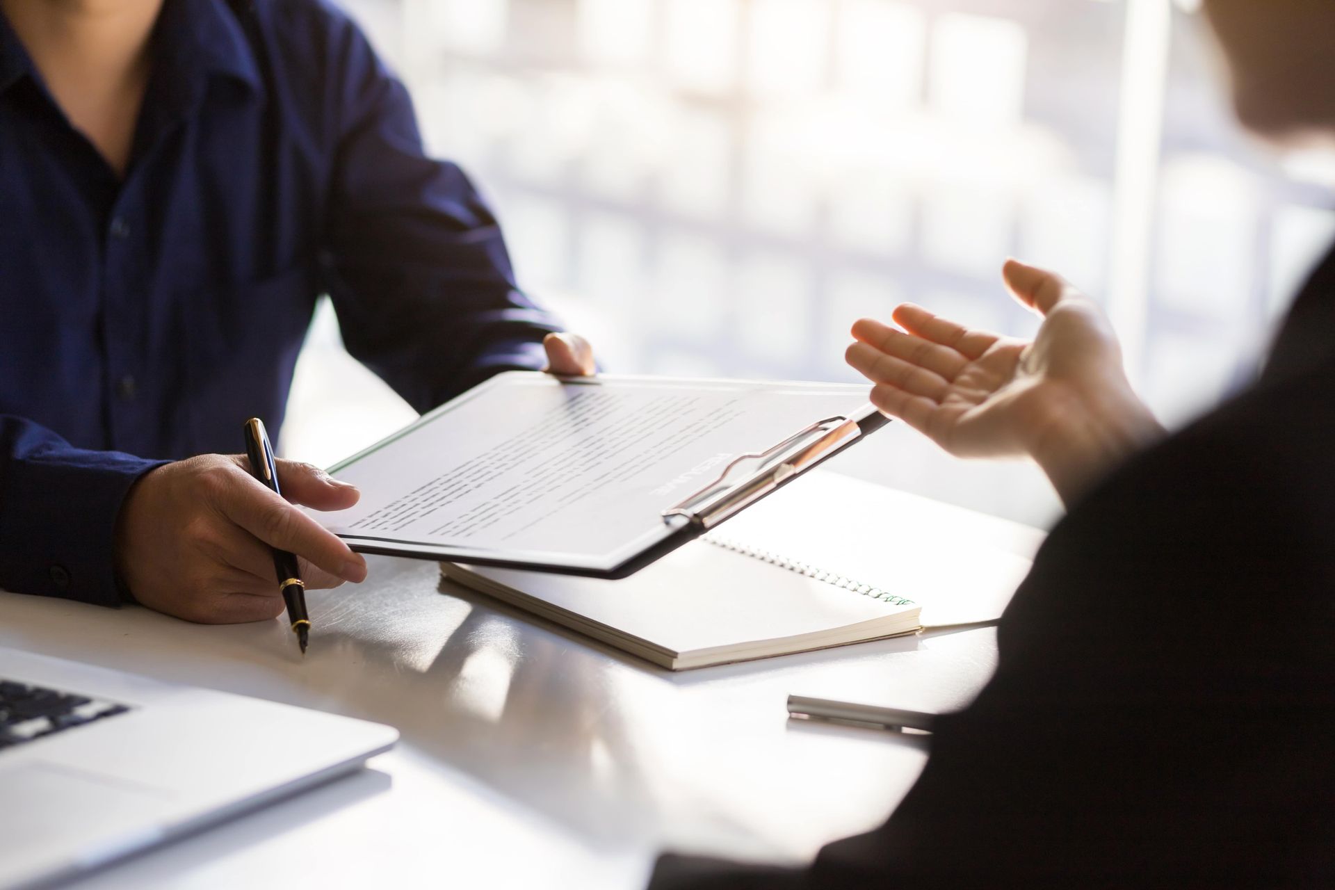 Two people discussing documents at a bright conference table, one taking notes with a pen
