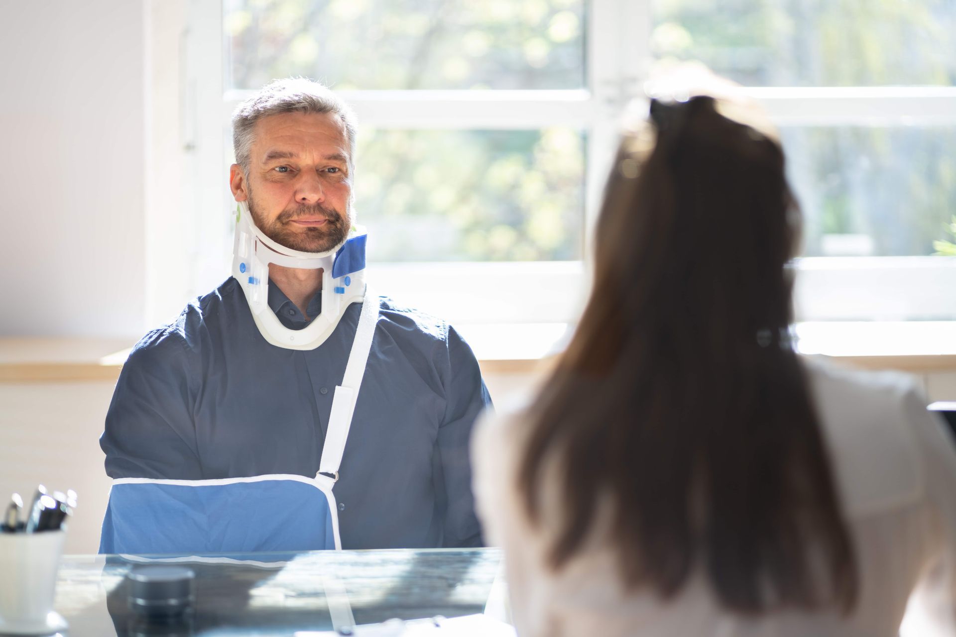 Person with a neck brace and arm sling speaking across a table in a bright office