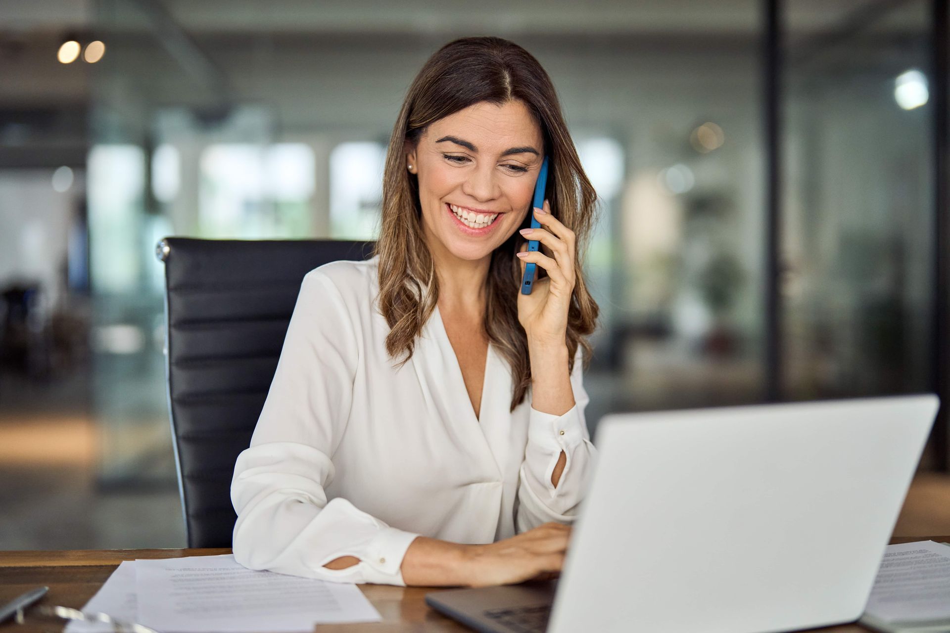 Woman smiling and talking on a phone while working on a laptop at an office desk
