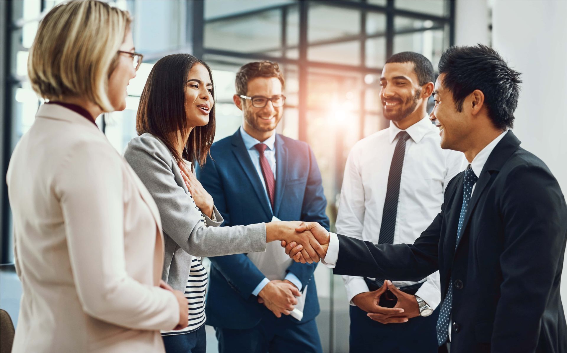 Business colleagues shaking hands in a bright office lobby, with others smiling nearby.