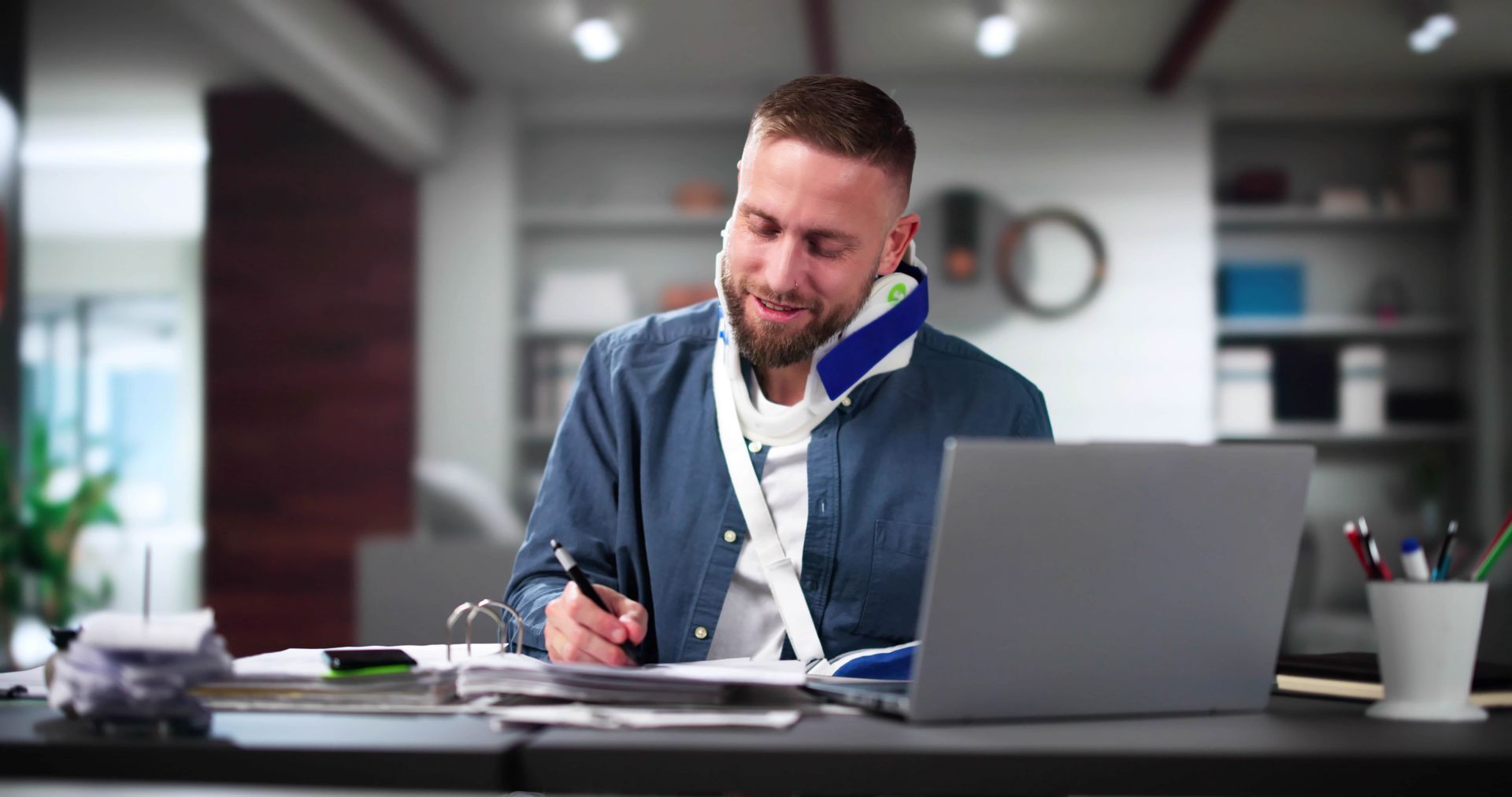 Person writing at a desk with a laptop in a modern office, wearing a blue jacket and looking focused.