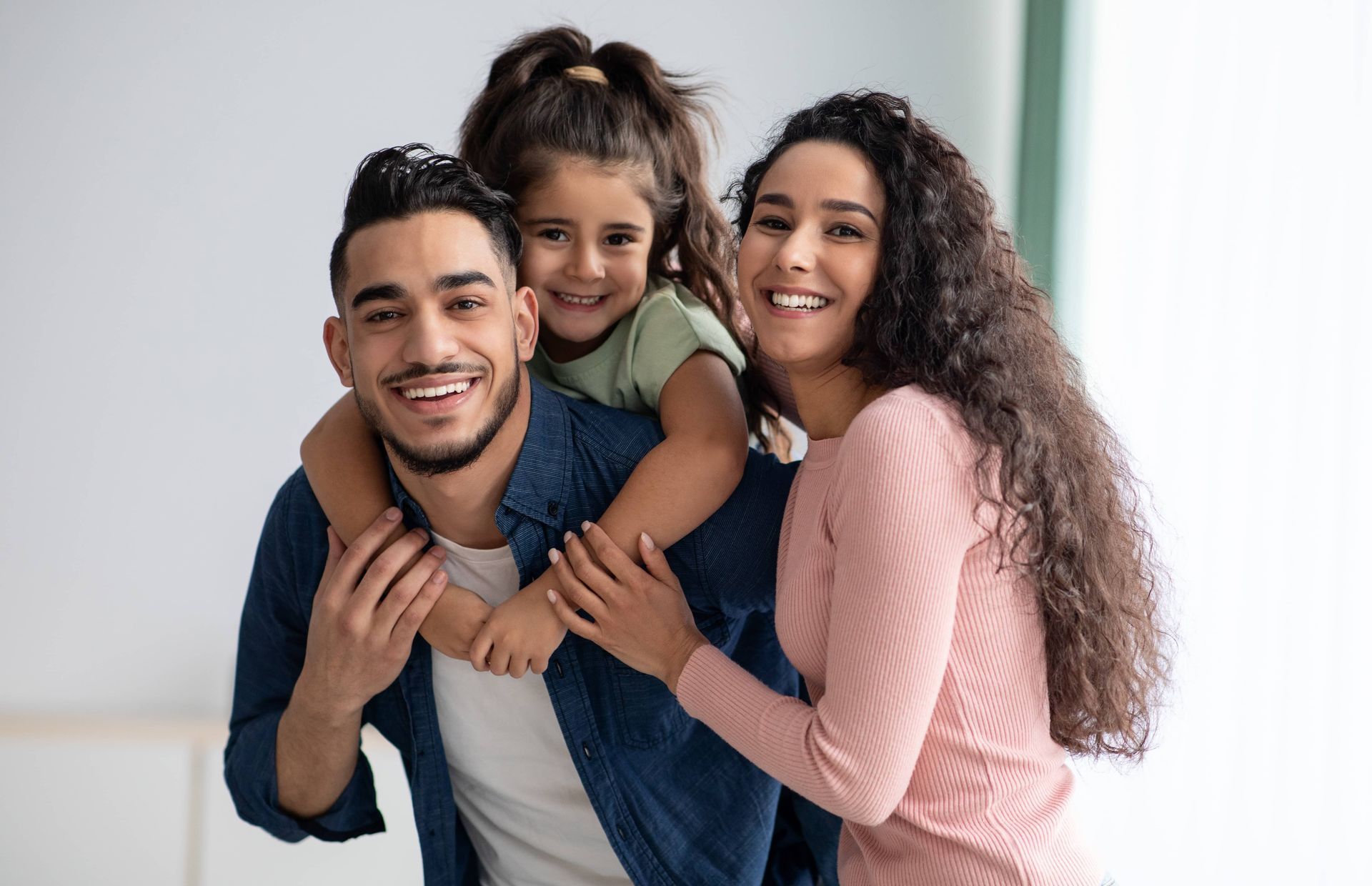 Smiling family of three posing together against a white background, with the child on the man's back.