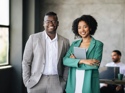 Two smiling coworkers in business attire standing in an office, one holding a tablet