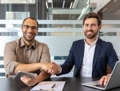 Two professionals shaking hands at a conference table, smiling in a bright office setting
