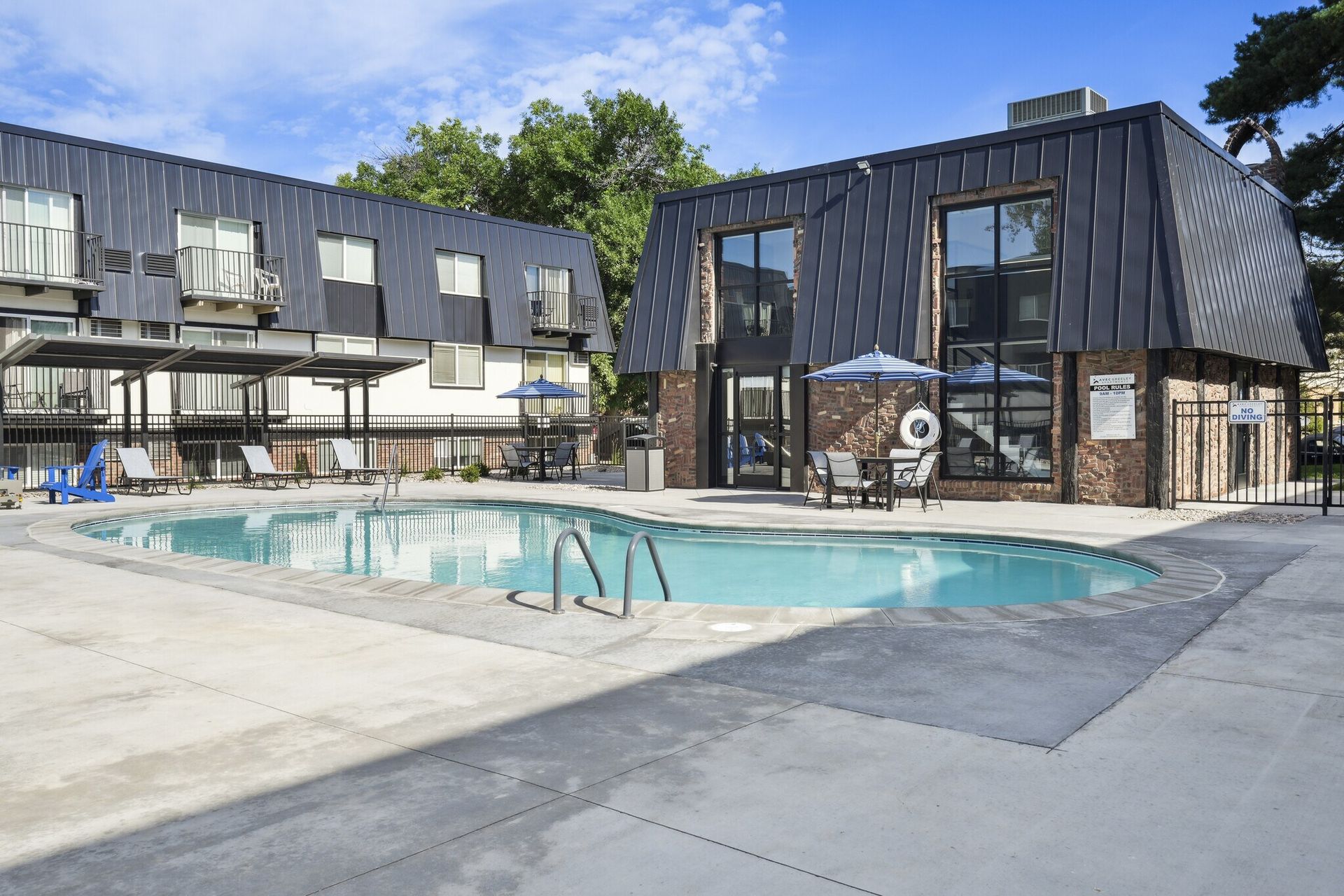A swimming pool sits in front of a multi-story apartment building with a dark, slanted roof and brick accents at Avec Greeley.