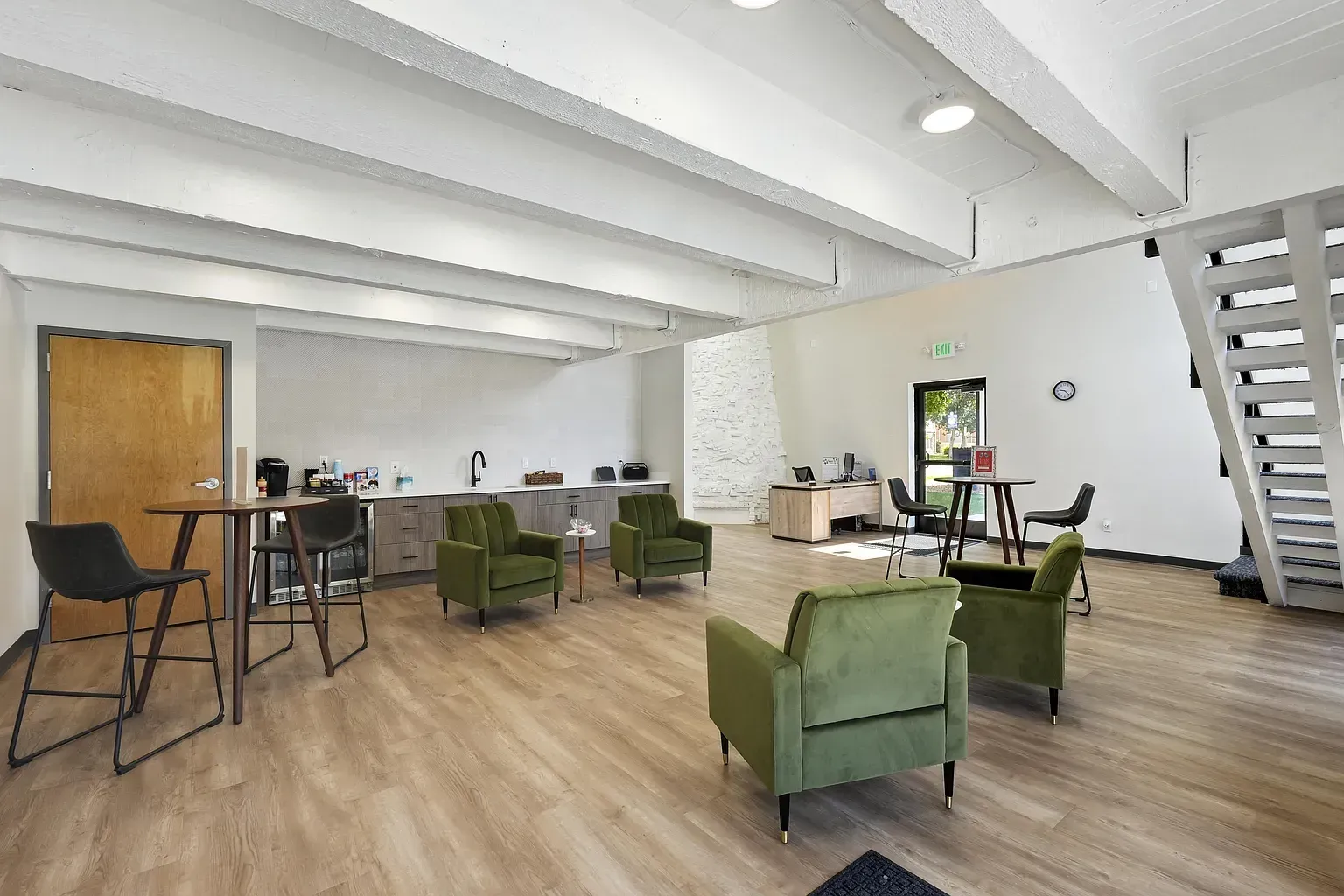 Modern office lounge with white walls, wooden floors, green velvet armchairs, a high-top table, and a staircase at Avec Greeley.
