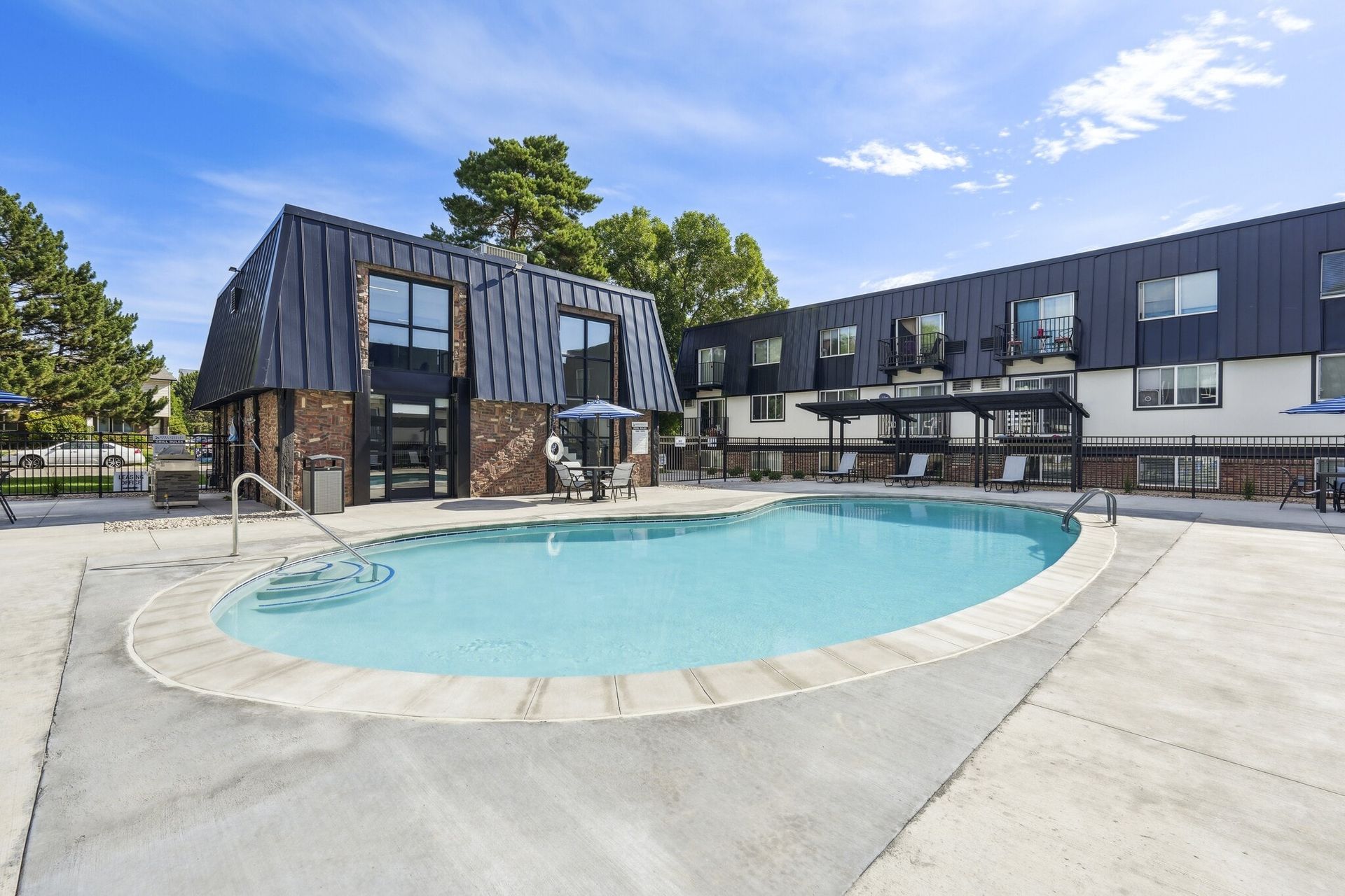 A kidney-shaped swimming pool in front of a multi-story hotel with black metal siding and a bright blue sky at Avec Greeley.
