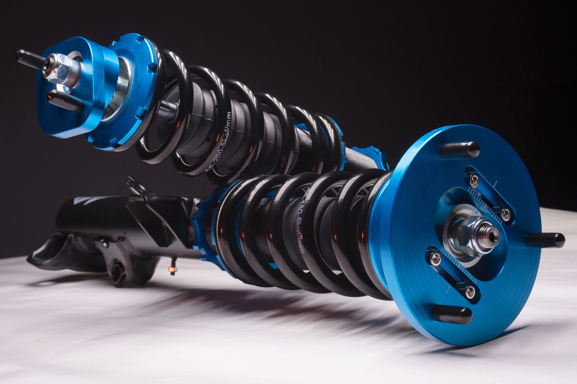 Sistema di sospensioni coilover blu e nero con molle nere, su una superficie bianca con sfondo scuro.