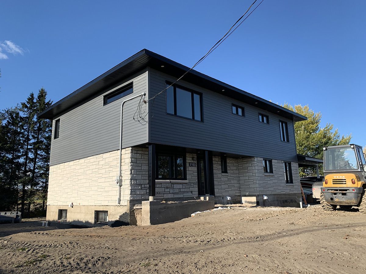 Maison à deux étages avec un bardage gris à l'étage et en pierre claire au rez-de-chaussée. Du matériel de chantier est stationné à proximité.