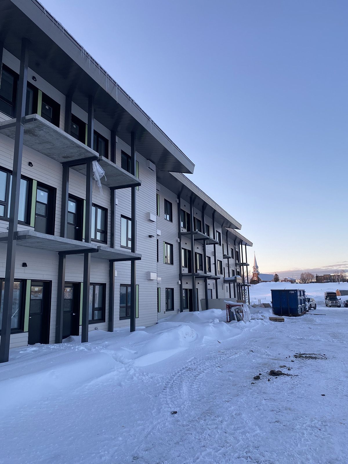 Un immeuble d'appartements de trois étages avec des balcons, un sol enneigé et un ciel bleu clair.