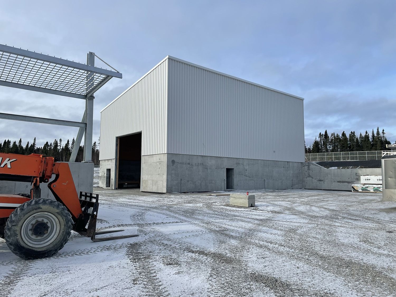 Chantier de construction avec un grand bâtiment blanc revêtu de métal sur des fondations en béton ; chariot élévateur au premier plan ; neige au sol.
