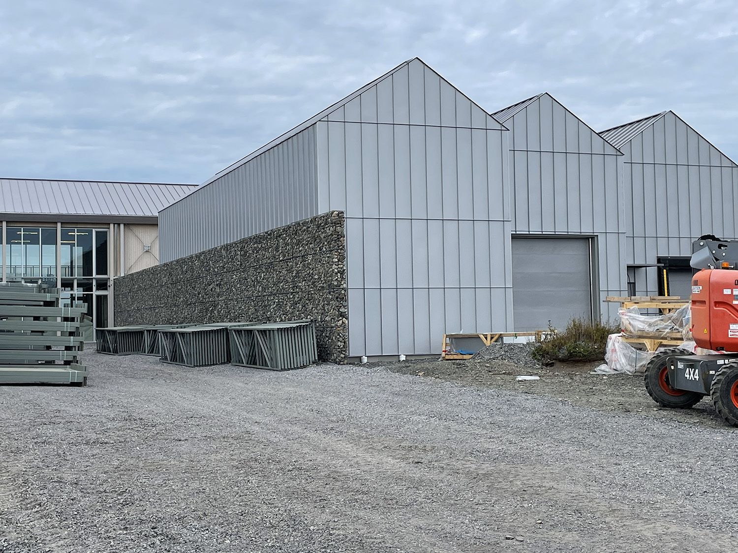 Extérieur d'un bâtiment gris avec un parking en gravier, un mur de pierres et des matériaux de construction.