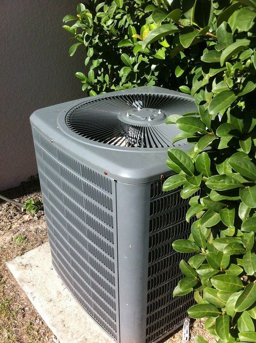 Outdoor air conditioner unit beside green bushes and a wall
