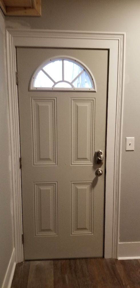 Tan door with arched window, hardware, and wood trim. Gray walls and brown wood floor.