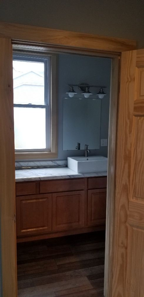 View of a bathroom interior through an open doorway. The room has a sink, vanity, mirror, and a window.