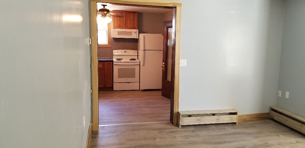 Interior view of a room with a doorway to a kitchen. Light blue walls, light wood floor. Kitchen has white appliances and wood cabinets.