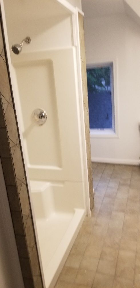 A white shower stall in a bathroom with beige tile flooring. A window is visible in the background.