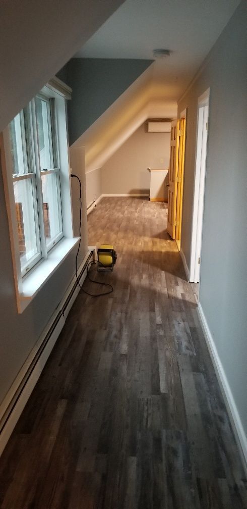 Narrow hallway with dark wood flooring, and sloped ceiling leading to a room with beige walls and a window.